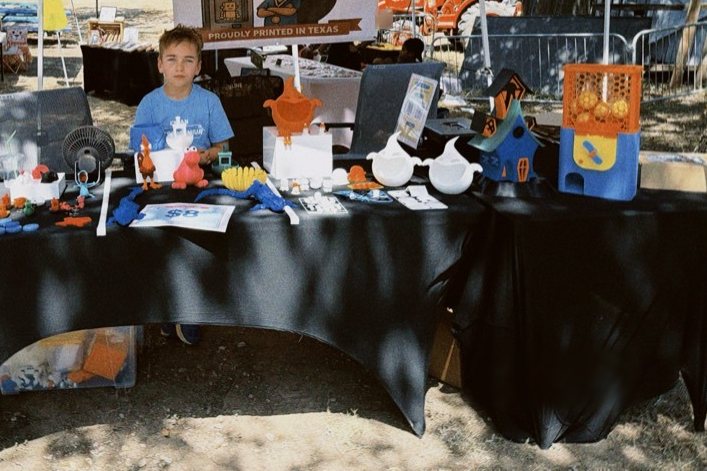 Joseph behind his market stall under a tent, the table covered in his 3D printed toys