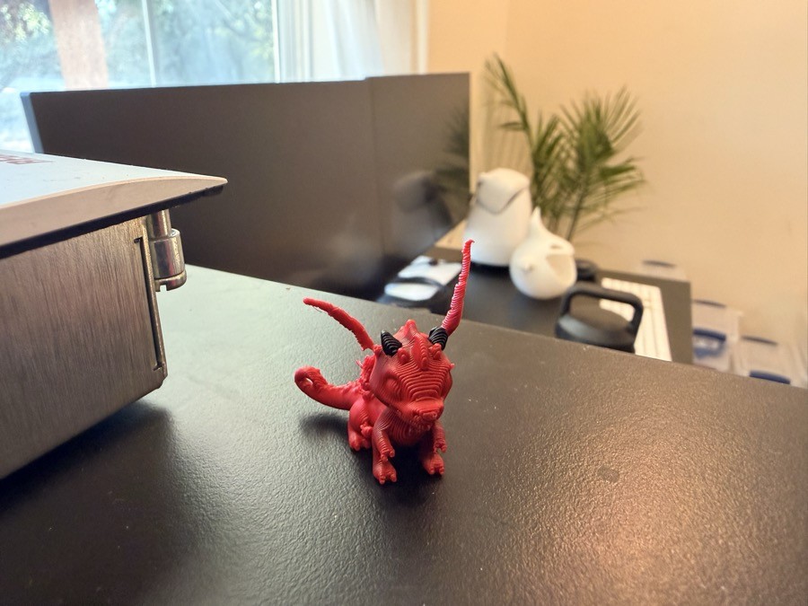 A small red 3D-printed dragon with black horns standing on a dark desk